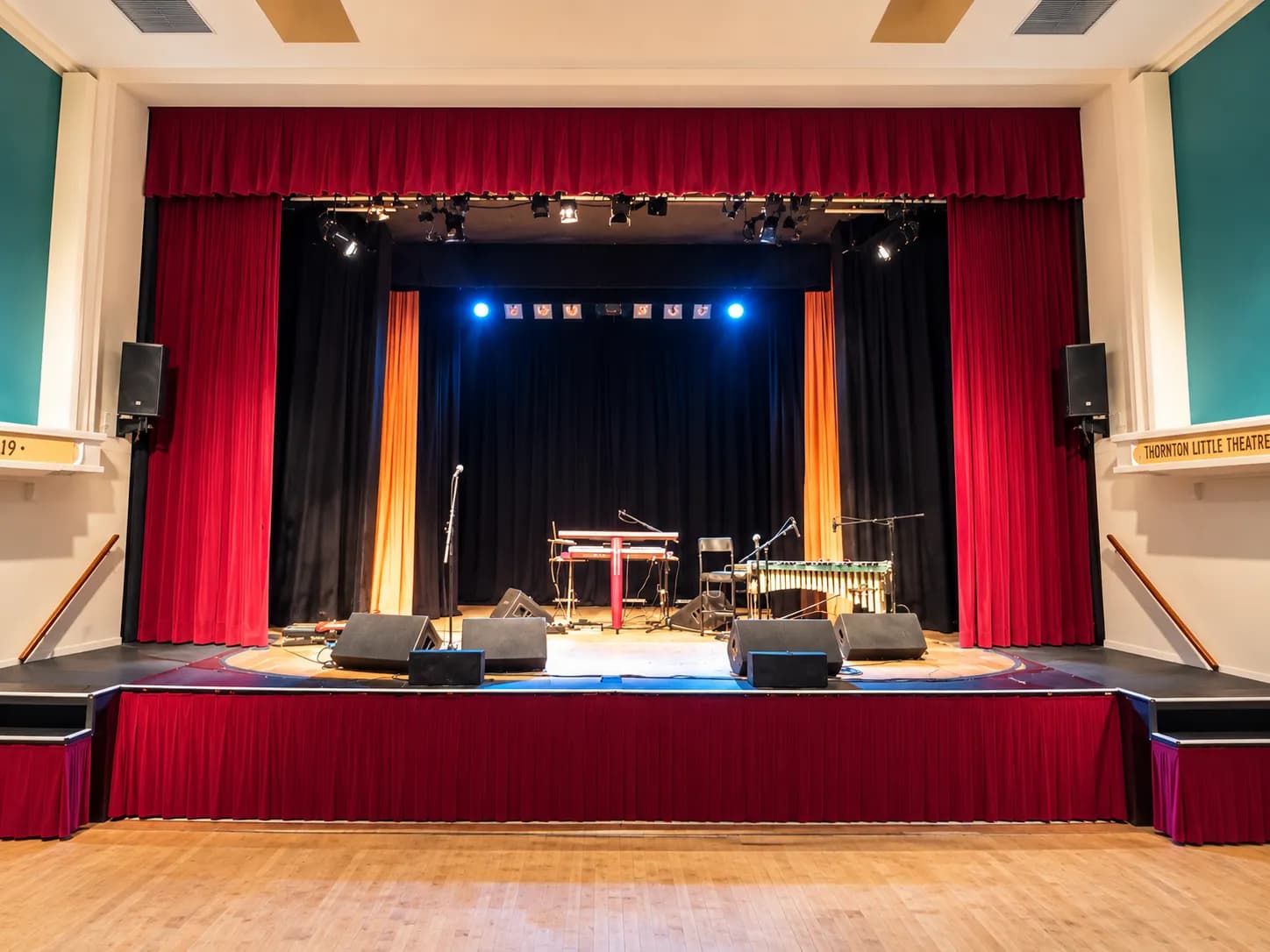 Thornton Little Theatre’s stage framed by red curtains, with blue lighting, microphones and musical instruments